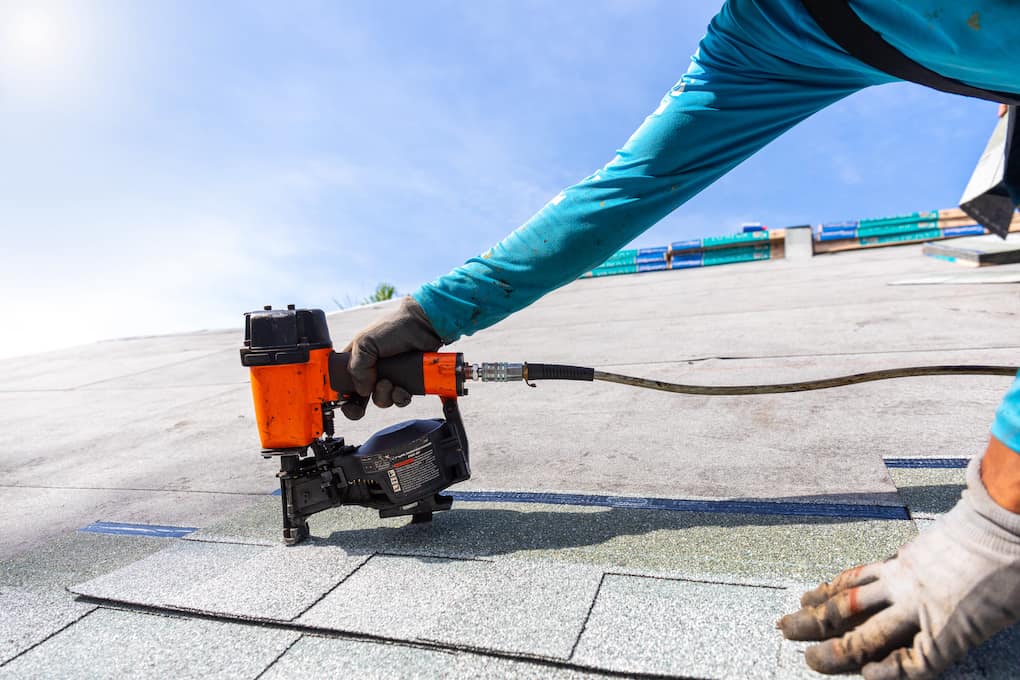 roofer installing roof shingles with pneumatic roofing nailer with blue sky background